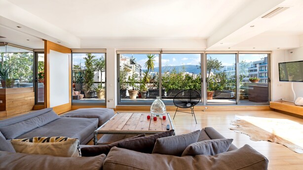 Uma sala de estar moderna com um sofá grande, uma mesa de centro de madeira e vista para uma varanda com plantas em vasos e para o horizonte da cidade.