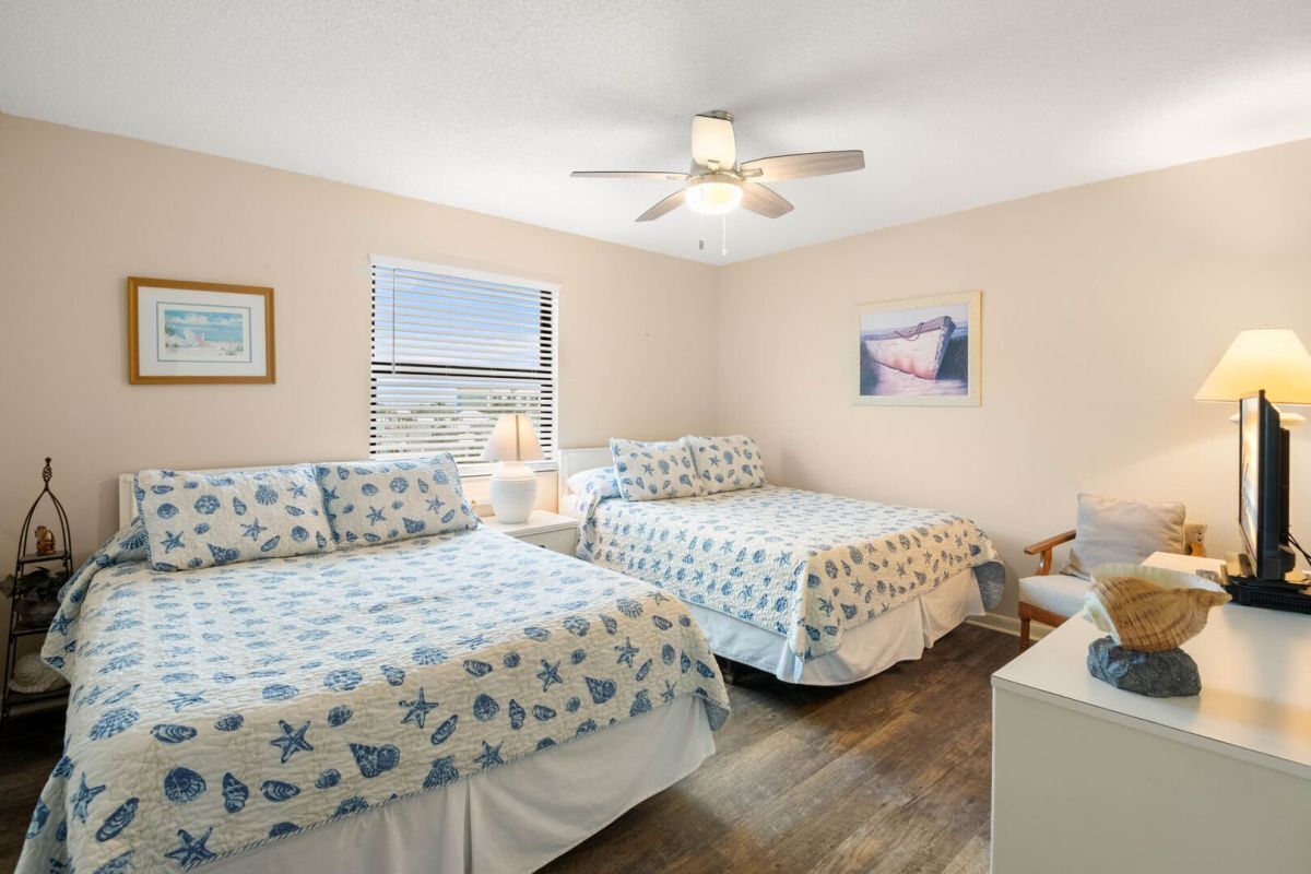 A bedroom with two beds, a ceiling fan, a window with blinds, a desk with a lamp, and a chair.