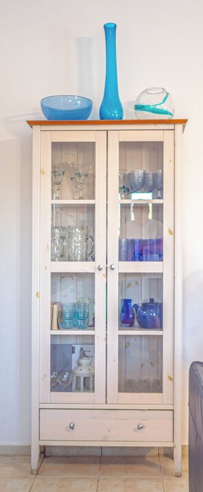 A white cabinet with glass doors containing various glassware and a blue vase on top.