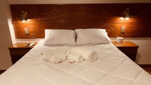 A neatly made bed with white linens and two rolled towels, a wooden headboard with geometric patterns, and bedside lamps. - Hotel Daylu Cusco (Cusco)