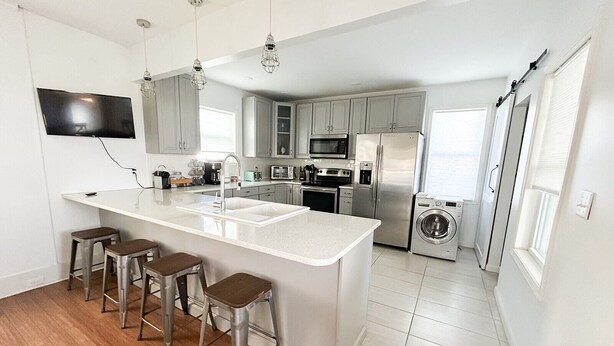 A modern kitchen with a central island, bar stools, stainless steel appliances, and a flat-screen TV mounted on the wall. - Sweet  Victorian Beach Cottage (Cape Charles)