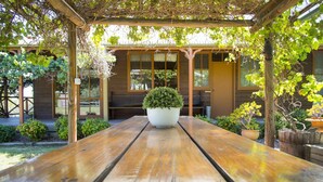 Ein Holztisch für ein Picknick mit einem Blumentopf in der Mitte, umgeben von einer Holzpergola und einem Gebäude mit großen Fenstern.