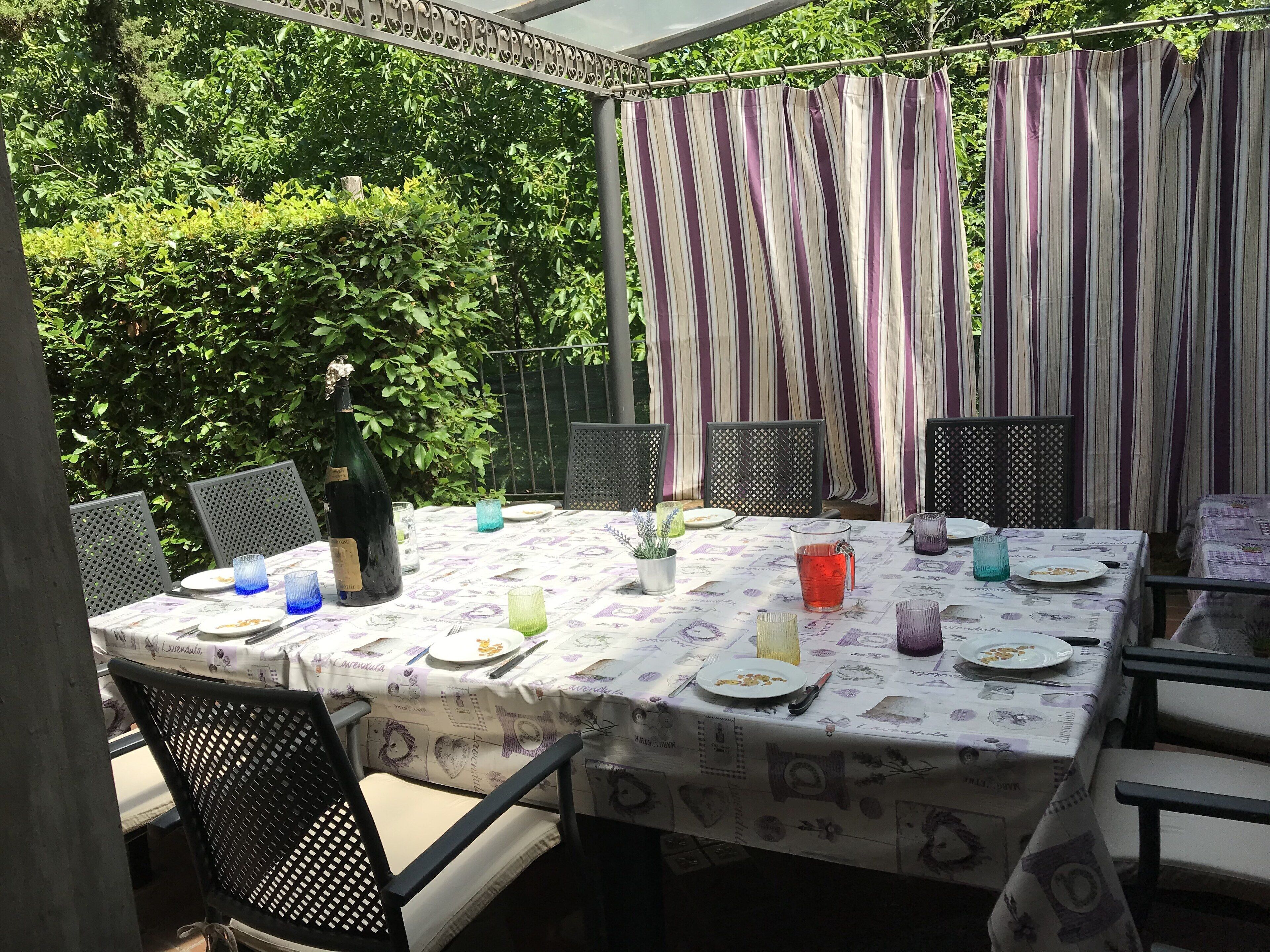Eine Terrasse im Freien mit einem Essplatz, umgeben von Stühlen und einem Vorhang als Hintergrund.