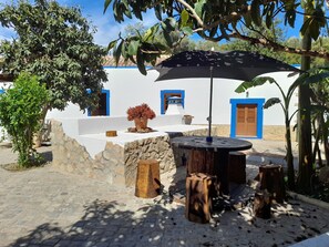A courtyard with a round wooden table and chairs, a black umbrella, and a potted plant. - Quinta Do Pomarinho 46328/AL  (Tavira)