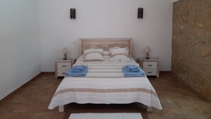 A bedroom with a bed, two bedside tables, and a stone wall. - Quinta Do Pomarinho 46328/AL  (Tavira)