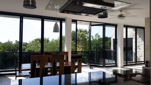 A modern dining area with a glass table and wooden chairs, large windows offering a view of trees, and a ceiling fan.