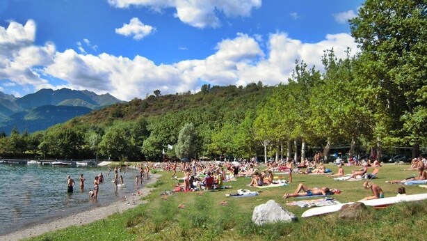 A crowded lakeside beach with people swimming and sunbathing, surrounded by lush greenery and mountains. - Cà Elsa B&B (Nuova Olonio)