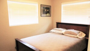 Une chambre à coucher avec un lit en bois, des murs de couleur beige et deux fenêtres équipées de stores.