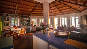 Ein geräumiges Zimmer mit hoher Holzbalkendecke, einem Essbereich mit Stühlen und Tisch, einem Bücherregal und einer Sitzecke mit Sofa und Kissen.