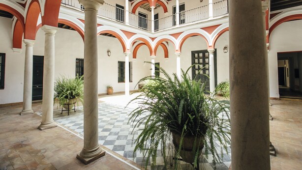 Un ampio cortile con pavimento a scacchi, circondato da colonne e archi, e piante in vaso.
