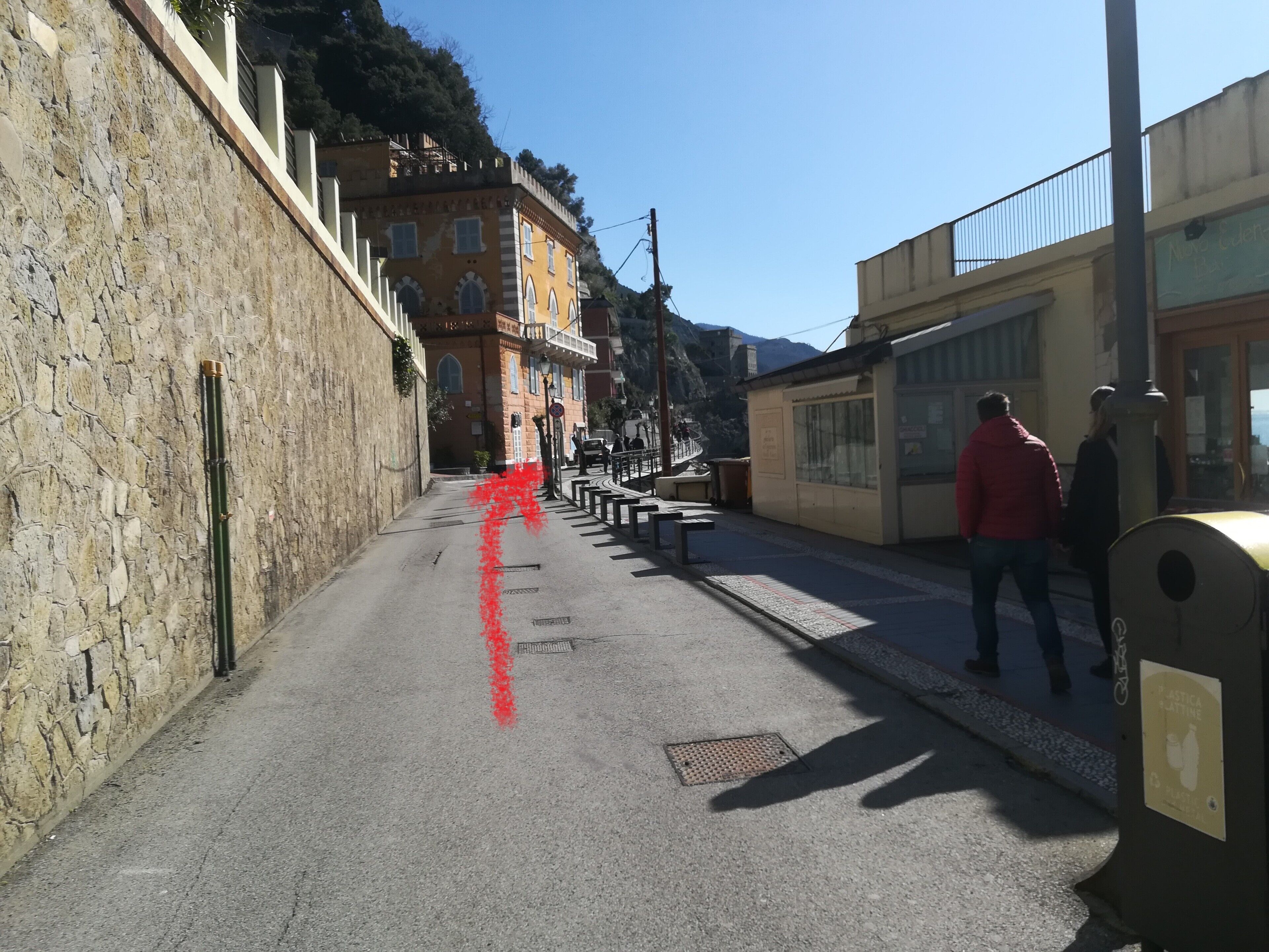 Een smalle straat met stenen muren aan één kant en gebouwen aan de andere kant. Er staan bankjes langs de stoeprand en er is een prullenbak aan de rechterkant.