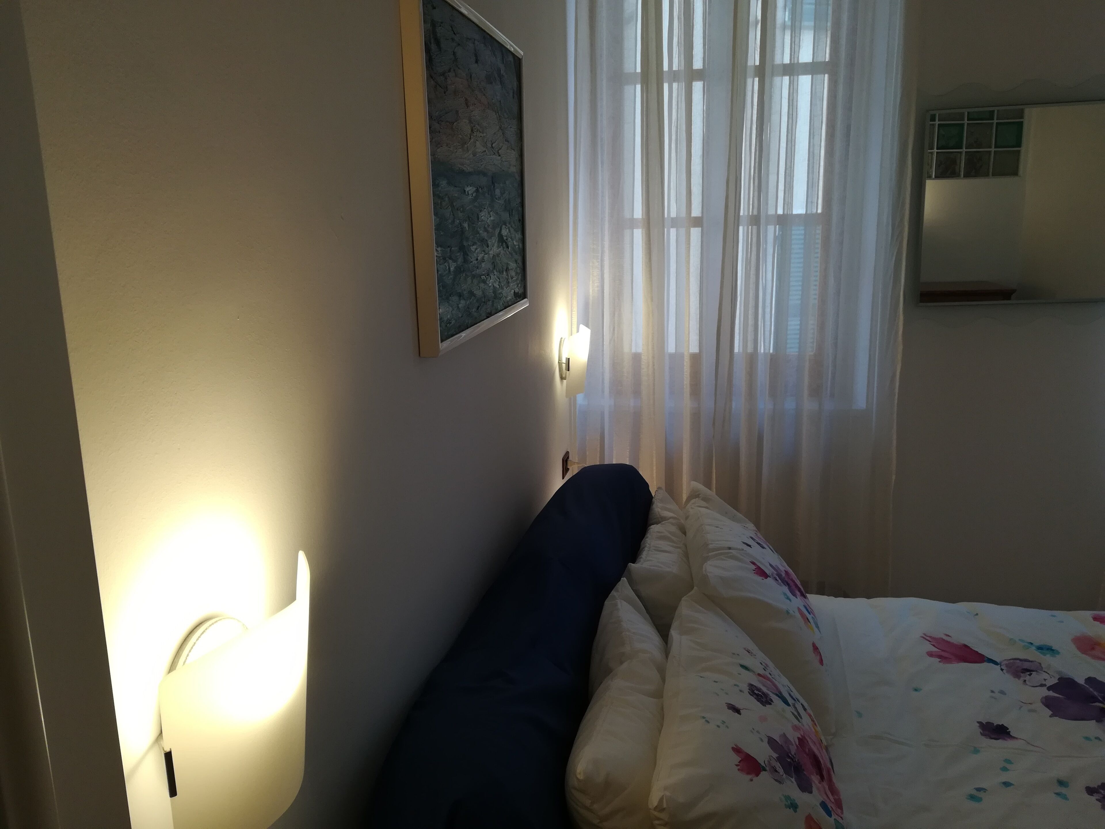 A bedroom with a bed, a wall-mounted light, a painting, and sheer curtains.