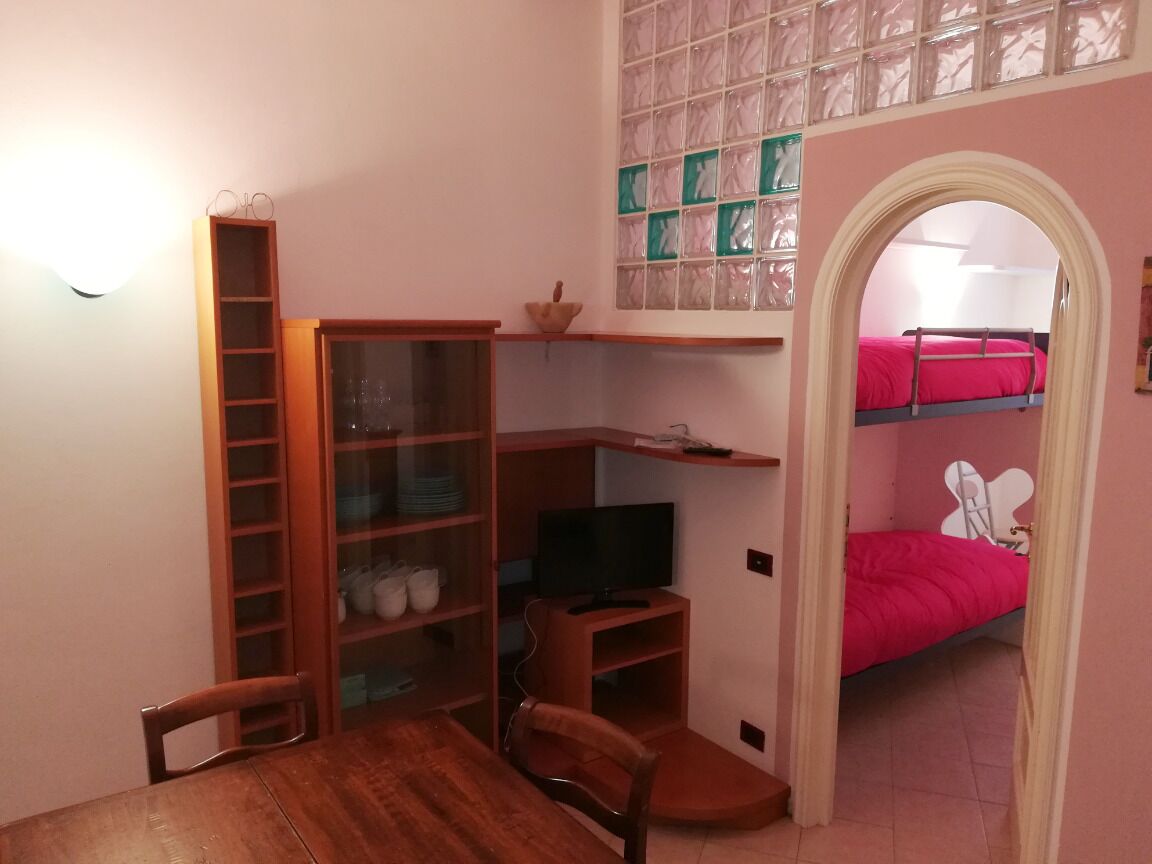 Een kleine kamer met een houten bureau, stoelen, een televisie, een stapelbed met een roze dekbedovertrek en een plank met diverse spullen.