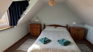 Un dormitorio con techo inclinado, una cama con edredón a cuadros, dos mesitas de noche, un aparador de madera, una lámpara colgante y una ventana con cortinas.