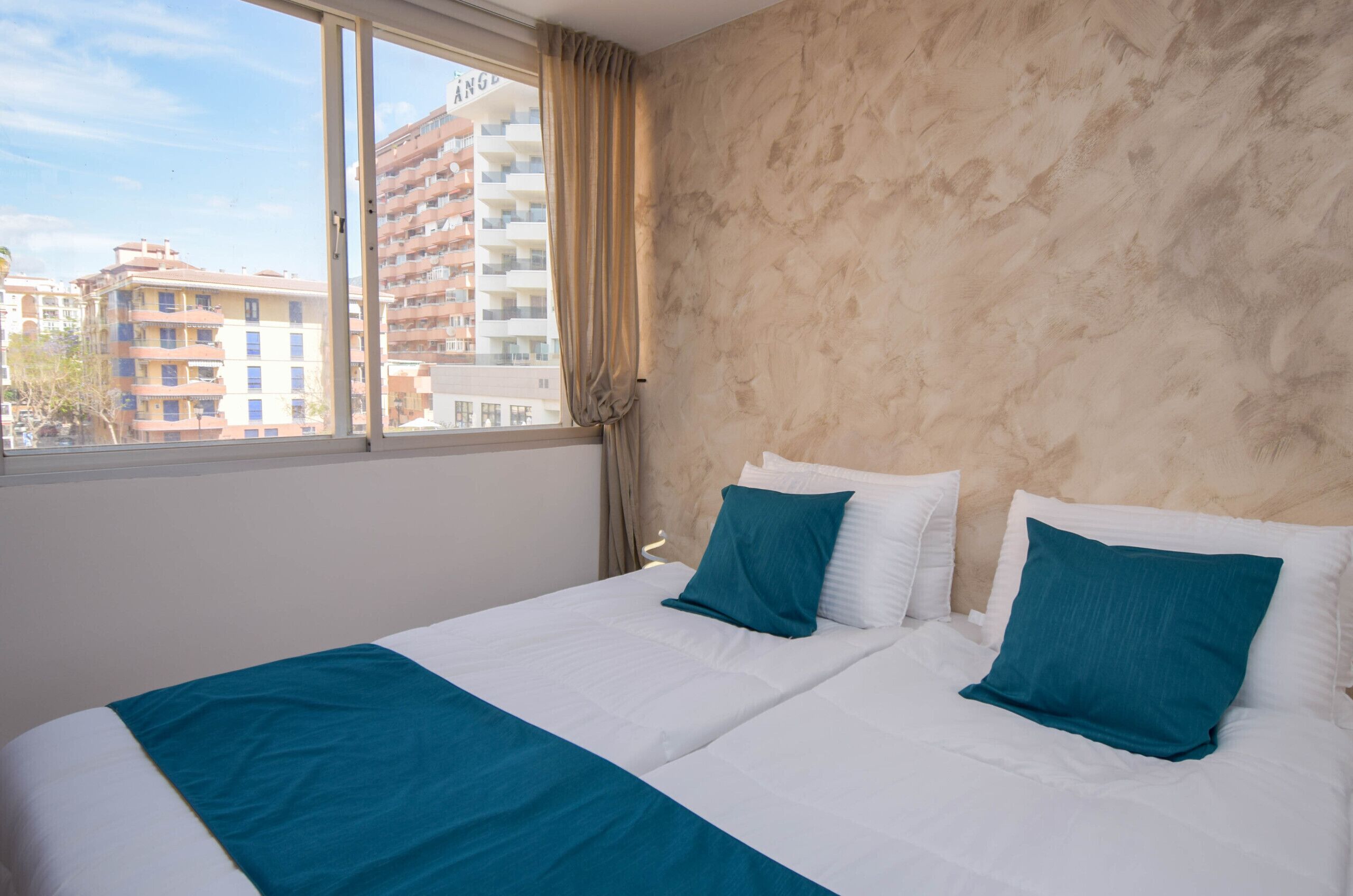 Een hotelkamer met een groot raam, twee bedden met wit en teal beddengoed, en een beige muur met textuur.