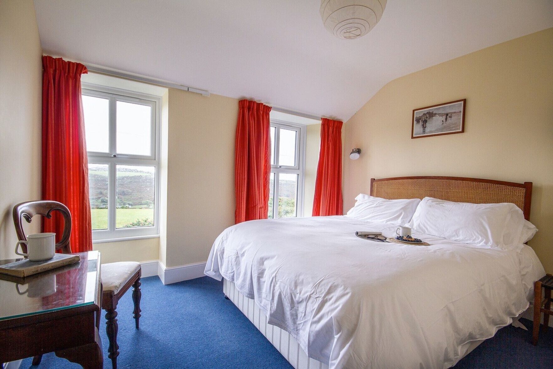 A bedroom with a large bed, a wooden headboard, a bedside table with a cup and a book, and a window with red curtains.