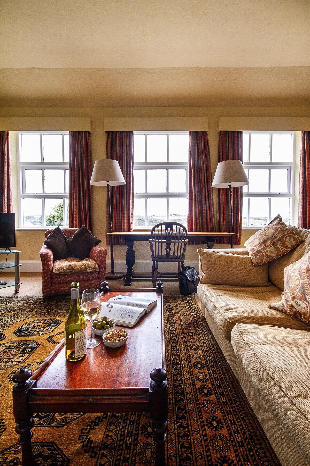 A living room with a sofa, armchair, wooden coffee table, and a patterned rug. There are two windows with curtains, a lamp, and a television.