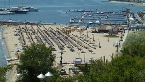 A beach with rows of sunbeds and umbrellas, a marina with boats, and a cityscape in the background.