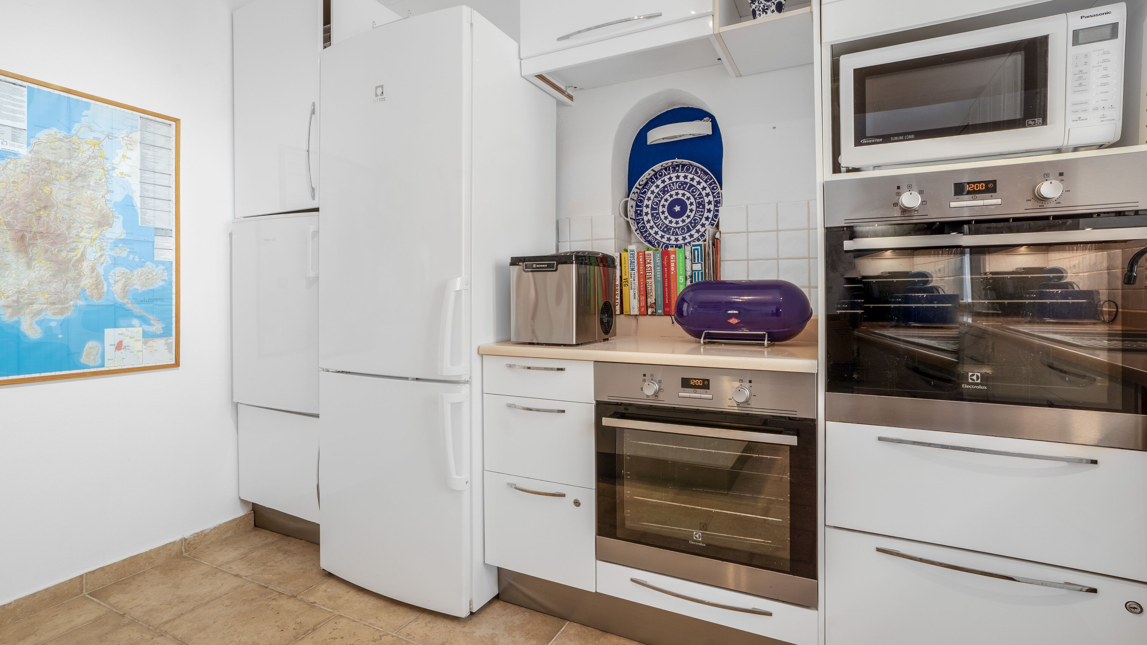 Eine Küche mit weißen Schränken, einem Kühlschrank, einer Mikrowelle, einem Backofen und einem Toaster. An der Wand hängt eine Karte und ein blauer dekorativer Teller.