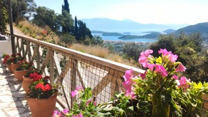 A balcony with potted flowers and a view of a coastal landscape with mountains. - A HOME FROM HOME! Large stone Villa with pool & stunning views - FREE meal out! (Lefkada)