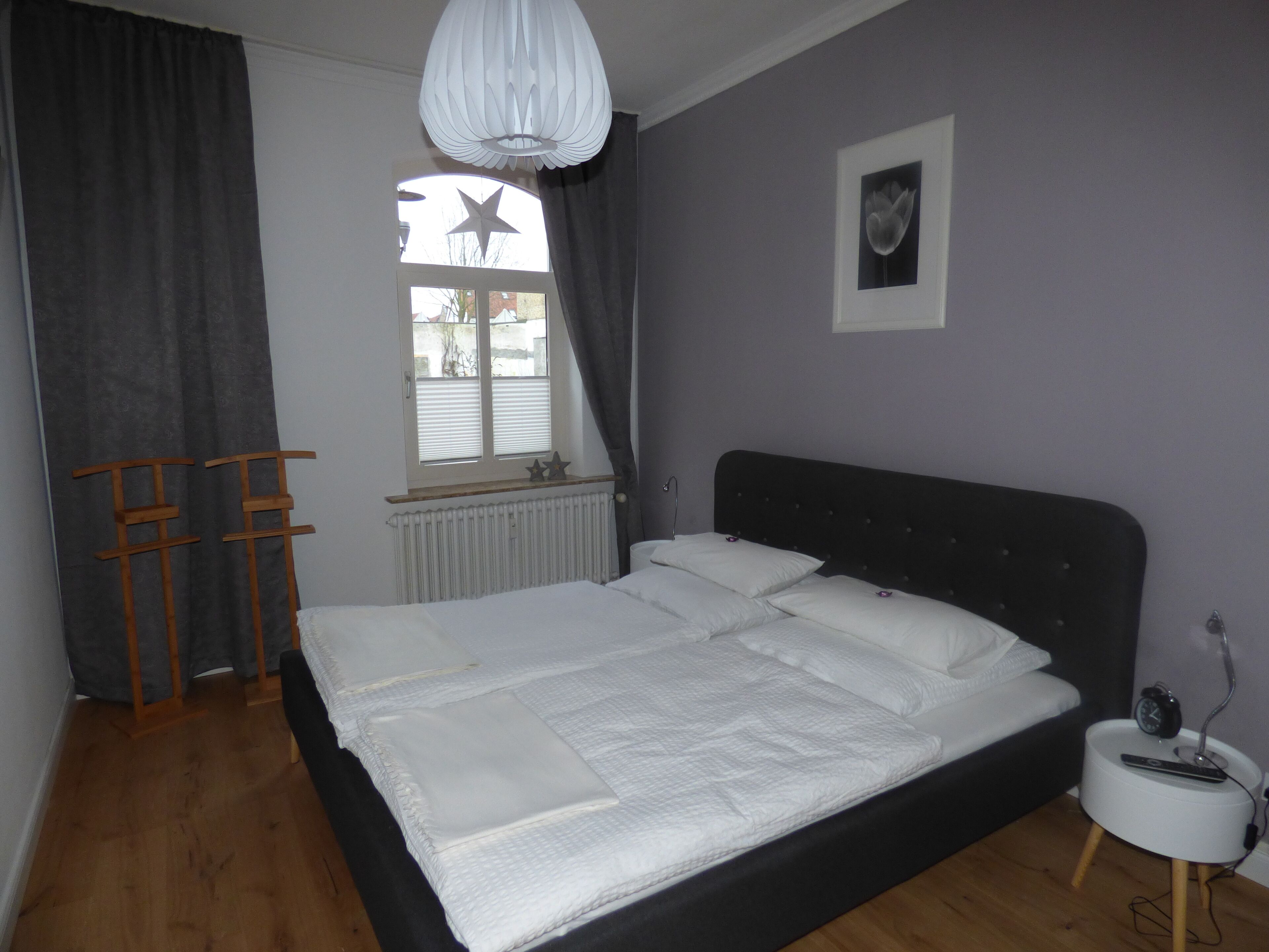 A bedroom with a bed, a window with curtains, a radiator, a bedside table with a lamp, and a wall-mounted picture.