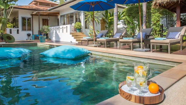 Zona junto a la piscina con sillones de descanso, sombrillas y una bandeja con bebidas y una naranja.