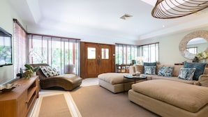 Amplia sala de estar con un sofá grande, una mesa de centro y un mueble de madera. Se observa un ventanal con persianas, una puerta y un ventilador de techo.