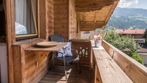 Ein Holzbalkon mit Tisch und Stuhl, ein Fenster mit Vorhängen und Blick auf Berge.