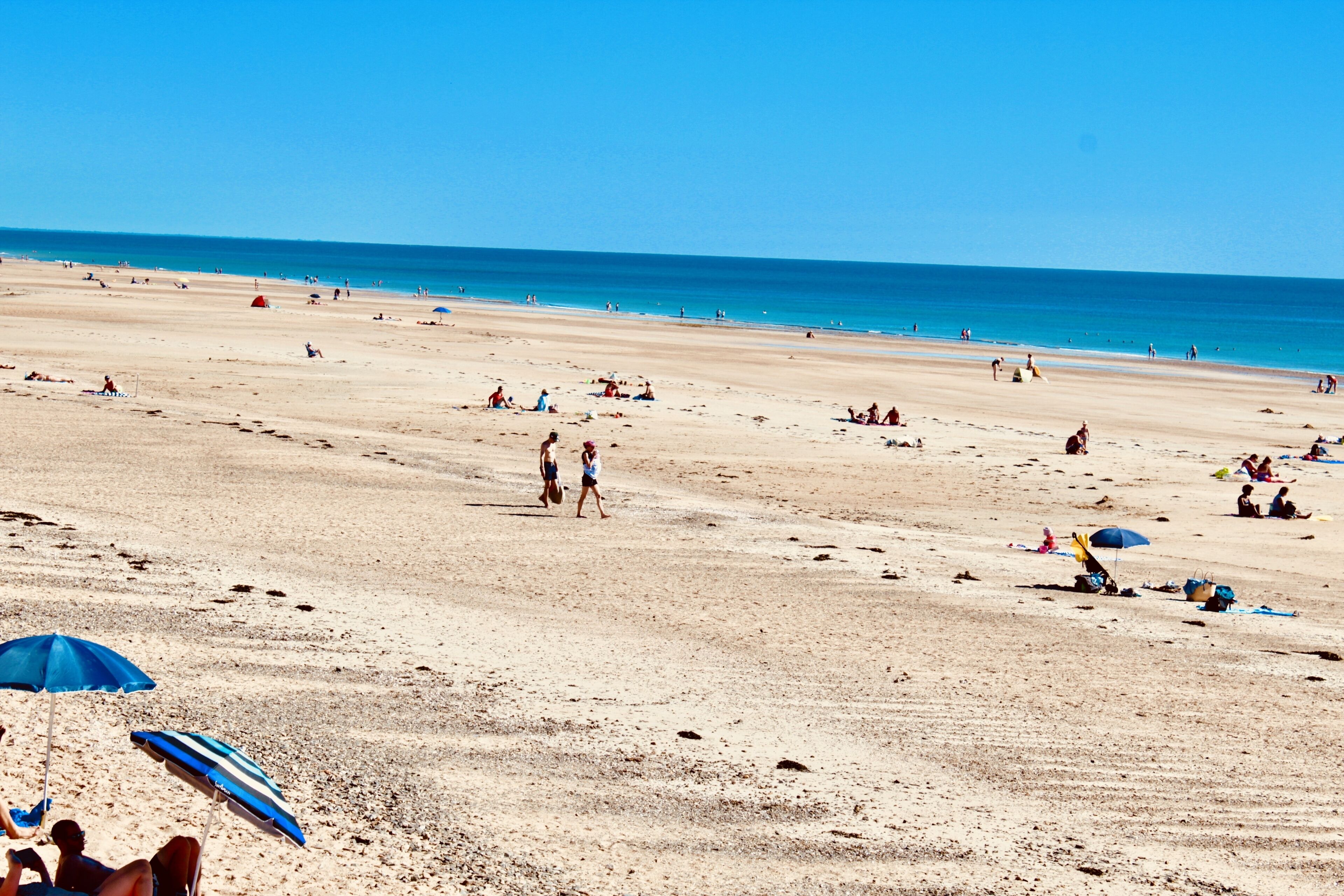 Een uitgestrekt zandstrand met mensen die wandelen en zitten, en een helderblauwe lucht.