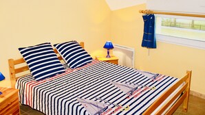 Een slaapkamer met een bed, gestreept beddengoed, een nachtkastje met een lamp, een raam met gordijnen en een aan de muur hangend schilderij.