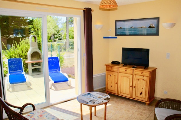A living room with a TV, wooden cabinets, a table with magazines, and a sliding glass door leading to a patio with blue chairs and a barbecue grill.