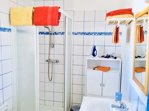 Een badkamer met een douche, wastafel en spiegel. Handdoeken hangen aan een rek en er ligt een strijkijzer op het aanrecht.
