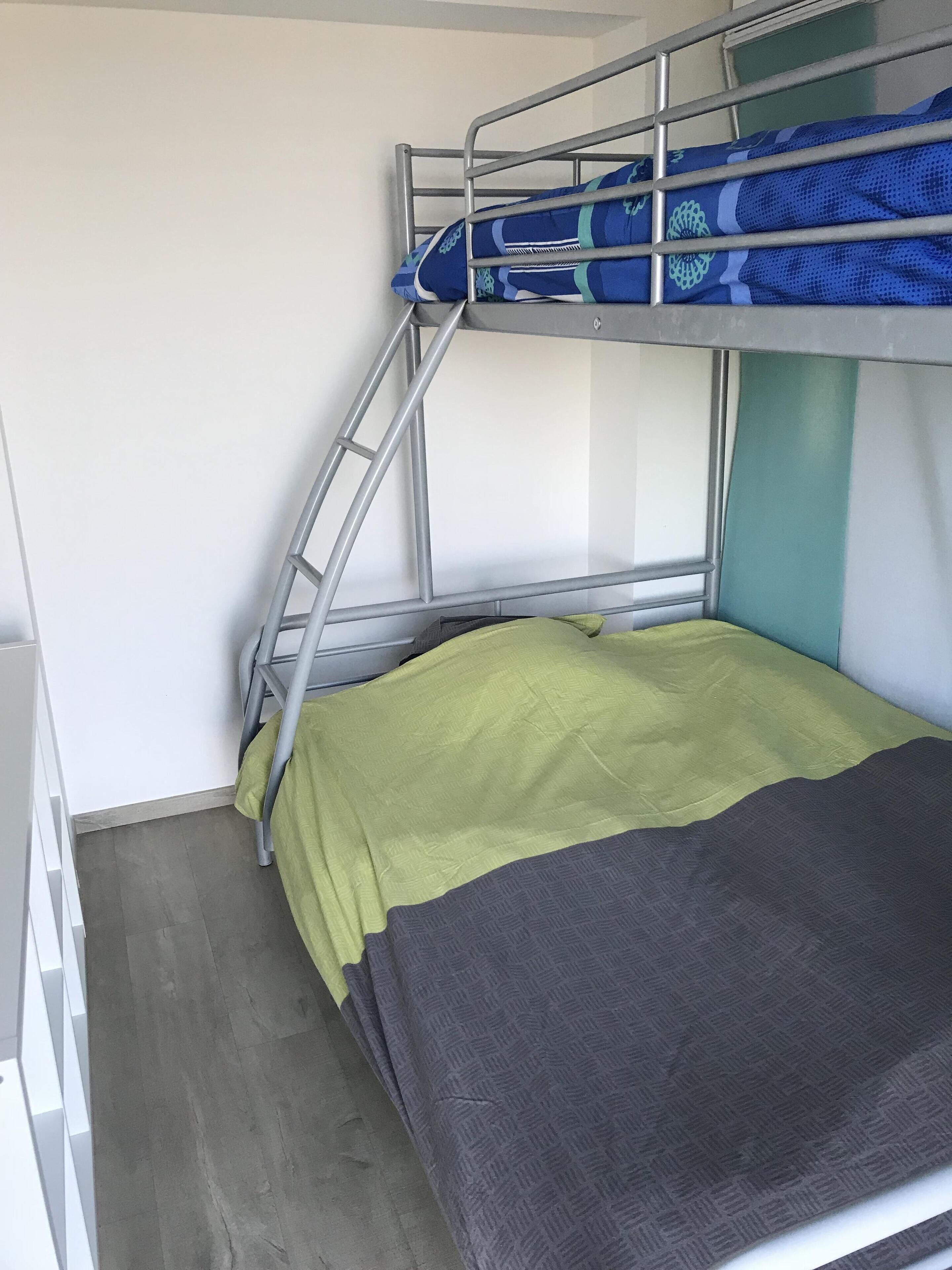 Un lit superposé avec une échelle, un matelas vert et un matelas gris, et un mur blanc.