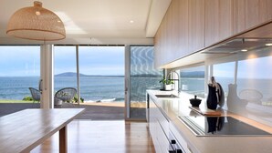 Een moderne keuken met een groot raam met uitzicht op zee, een eethoek met een houten tafel en een hanglamp.