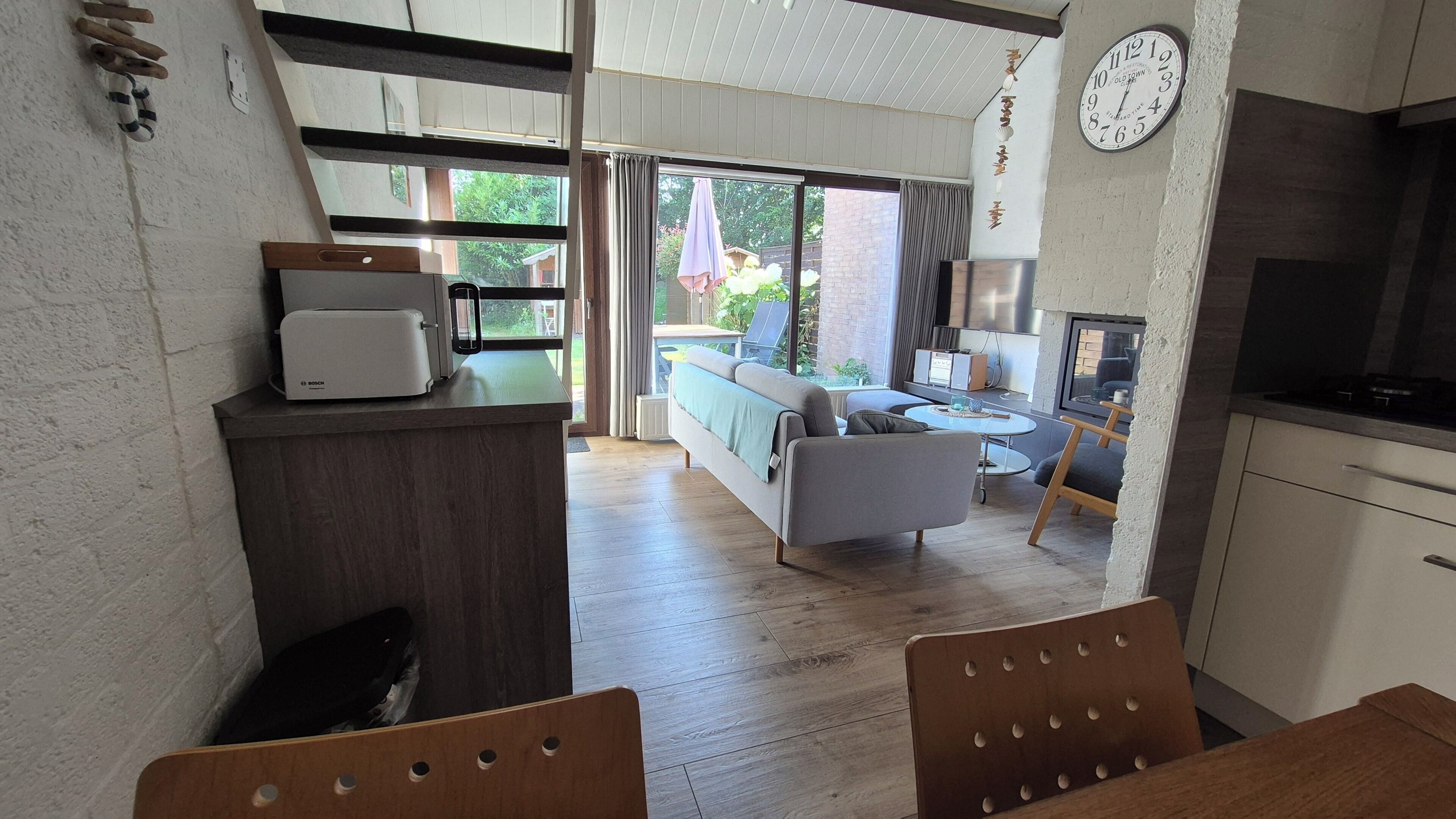 A modern kitchen with a Bosch refrigerator, a dining area with wooden chairs, a sofa, and a view of the garden through large windows.