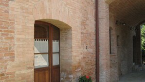 Un bâtiment en briques avec une porte en bois, une petite fenêtre ornée d’un treillis et deux plantes en pot fleuries.