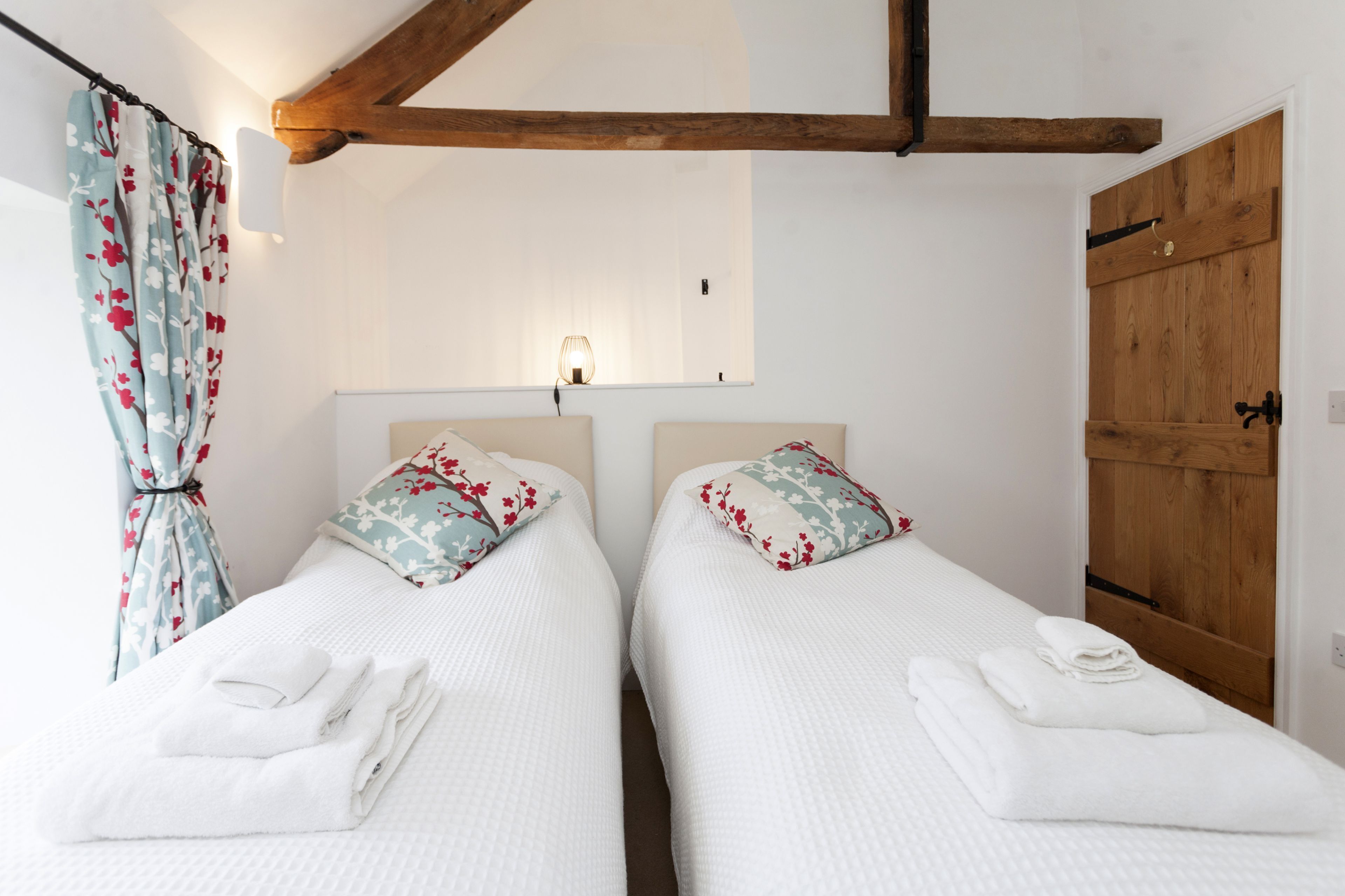 A room with two beds, each with a pillow and a folded towel. There is a wooden door and a window with curtains.