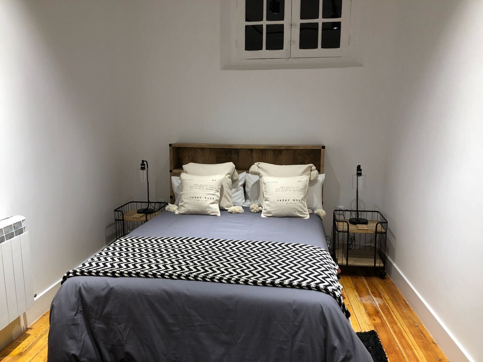 A bedroom with a bed, two bedside tables, a window, and a radiator.