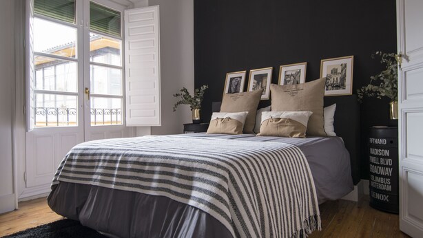 Een slaapkamer met een bed, een raam met luiken, een zwarte muur en lijstjes met foto's aan de muur.