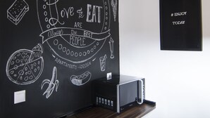 Een kamer met een prikbordmuur met tekeningen en tekst over eten, een houten bureau met een computer en een zwarte speaker aan de muur.