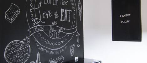 Una habitación con un muro de pizarra con dibujos y texto sobre comida, un escritorio de madera con una computadora y un altavoz negro en la pared.