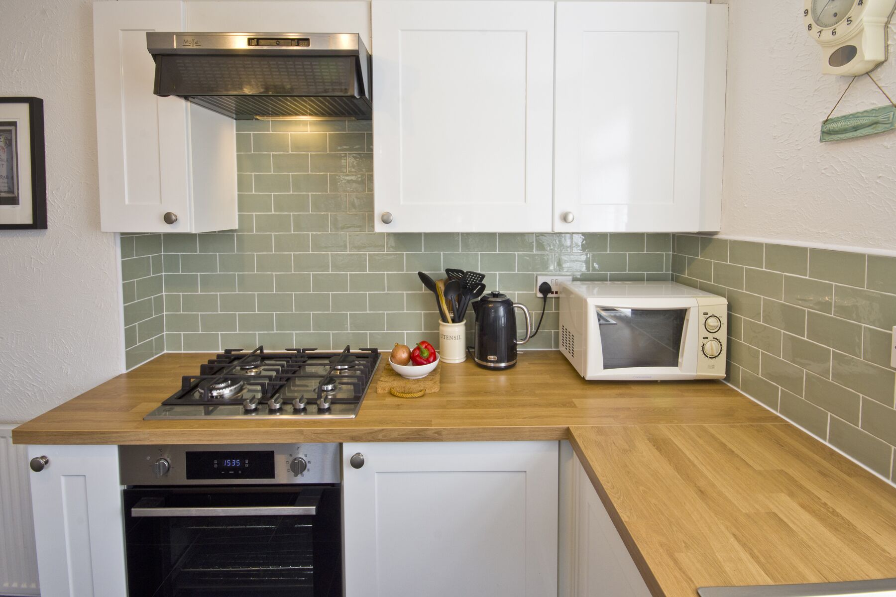 Een keuken met witte kastjes, een gasfornuis, een oven, een magnetron en een groene tegeltjeswand.
