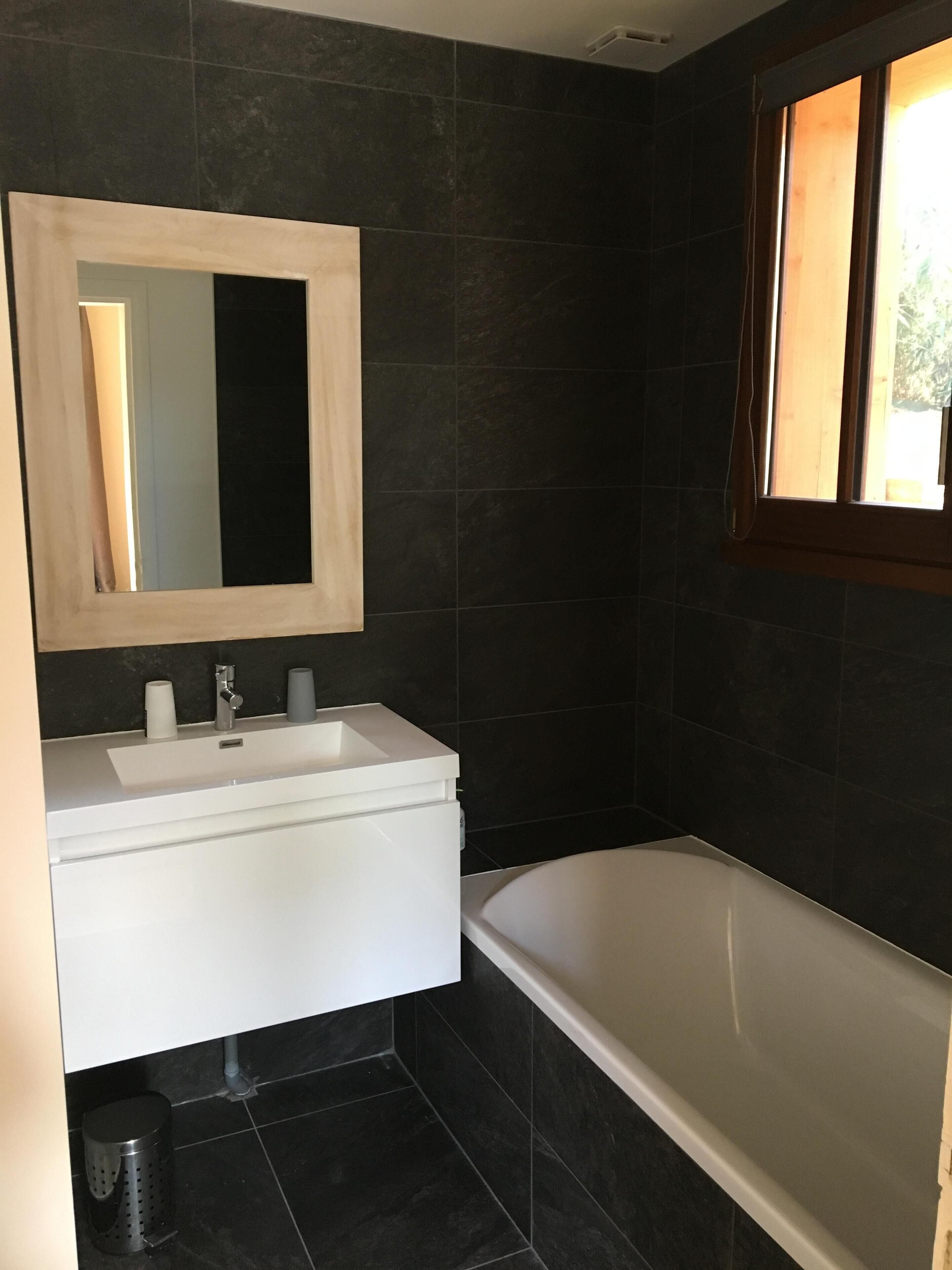 A modern bathroom with dark tiles, a white sink, a mirror, and a bathtub.