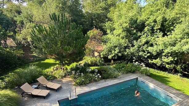 A rectangular swimming pool with a wooden deck, surrounded by lush greenery and trees.