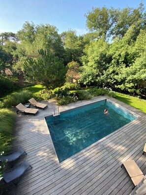 A rectangular swimming pool with a wooden deck, surrounded by lush greenery and trees.