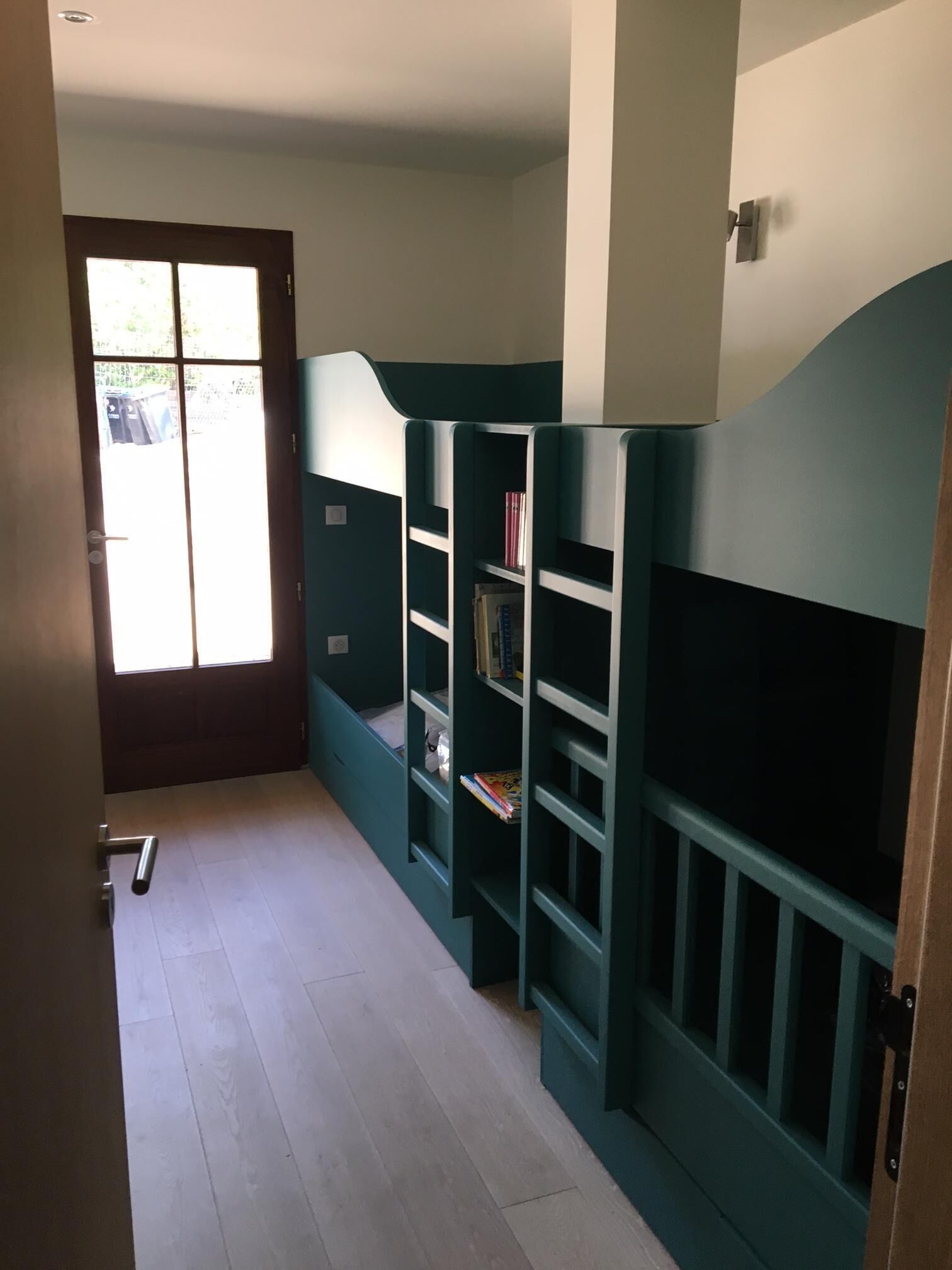 A room with a door leading to an outdoor area, a wooden floor, and green bunk beds with bookshelves.