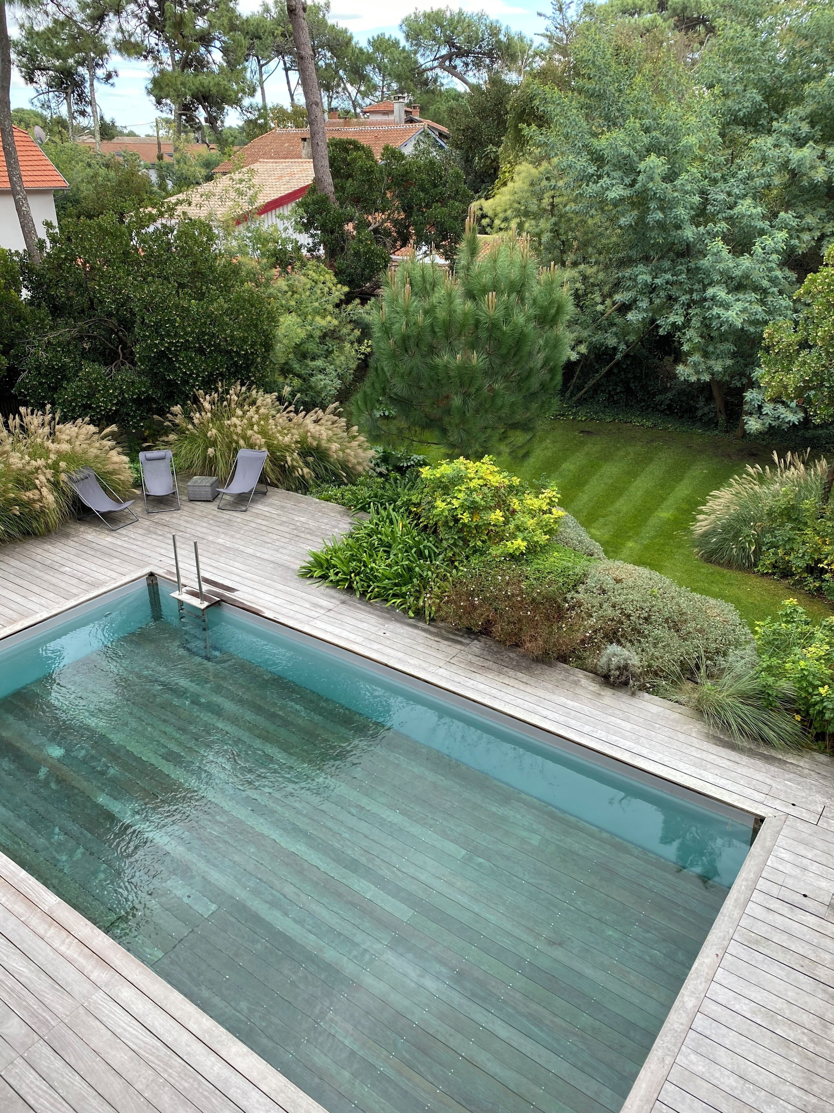 A rectangular swimming pool with a wooden deck surrounding it, three lounge chairs, and various shrubs and trees in the background.