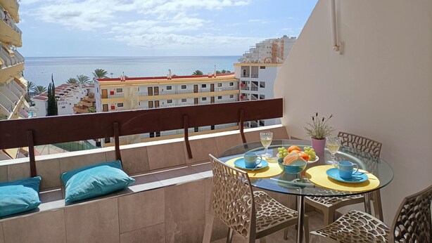 Un balcon avec une table en verre dressée pour le dîner, offrant une vue sur la côte, avec des bâtiments et la mer.