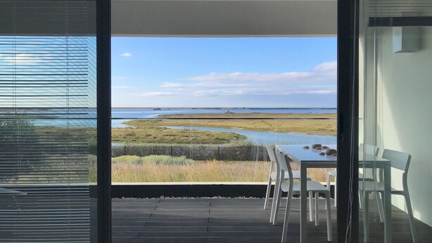 Ein Zimmer mit einer großen Glasdoor, die auf eine Terrasse mit Tisch und Stühlen führt, mit Blick auf eine Küstenlandschaft mit Wasser und Rasenflächen.