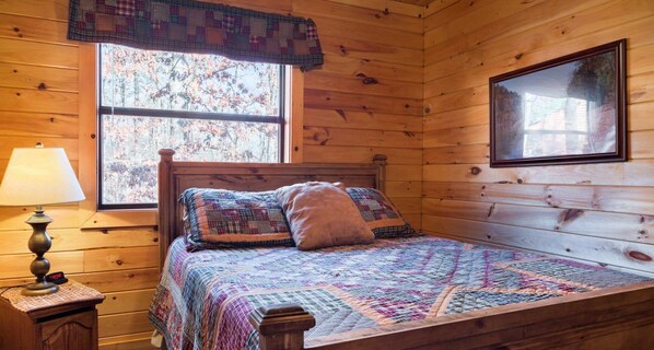 A wooden cabin room with a bed, a window, a lamp, and a framed picture on the wall.